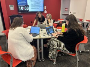 Four people seated around a small table in a classroom with red and white walls, each working on a laptop. A large screen on the wall displays a slide titled ten minutes decluttering or fixing content, with bullet points about accessibility tasks and a visible ten‑minute timer.