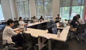 Photo of people seated around a U‑shaped arrangement of white desks in a modern classroom, each working on a laptop. Items such as water bottles, papers, and name tags are on the tables. Large windows provide natural light and a view of trees and a walkway outside.