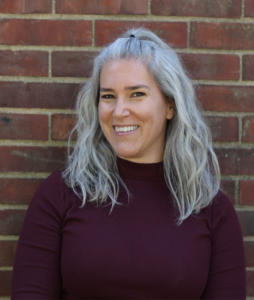 A woman with long gray hair and a dark colored turtle neck shirt, standing against a brick wall, smiling.