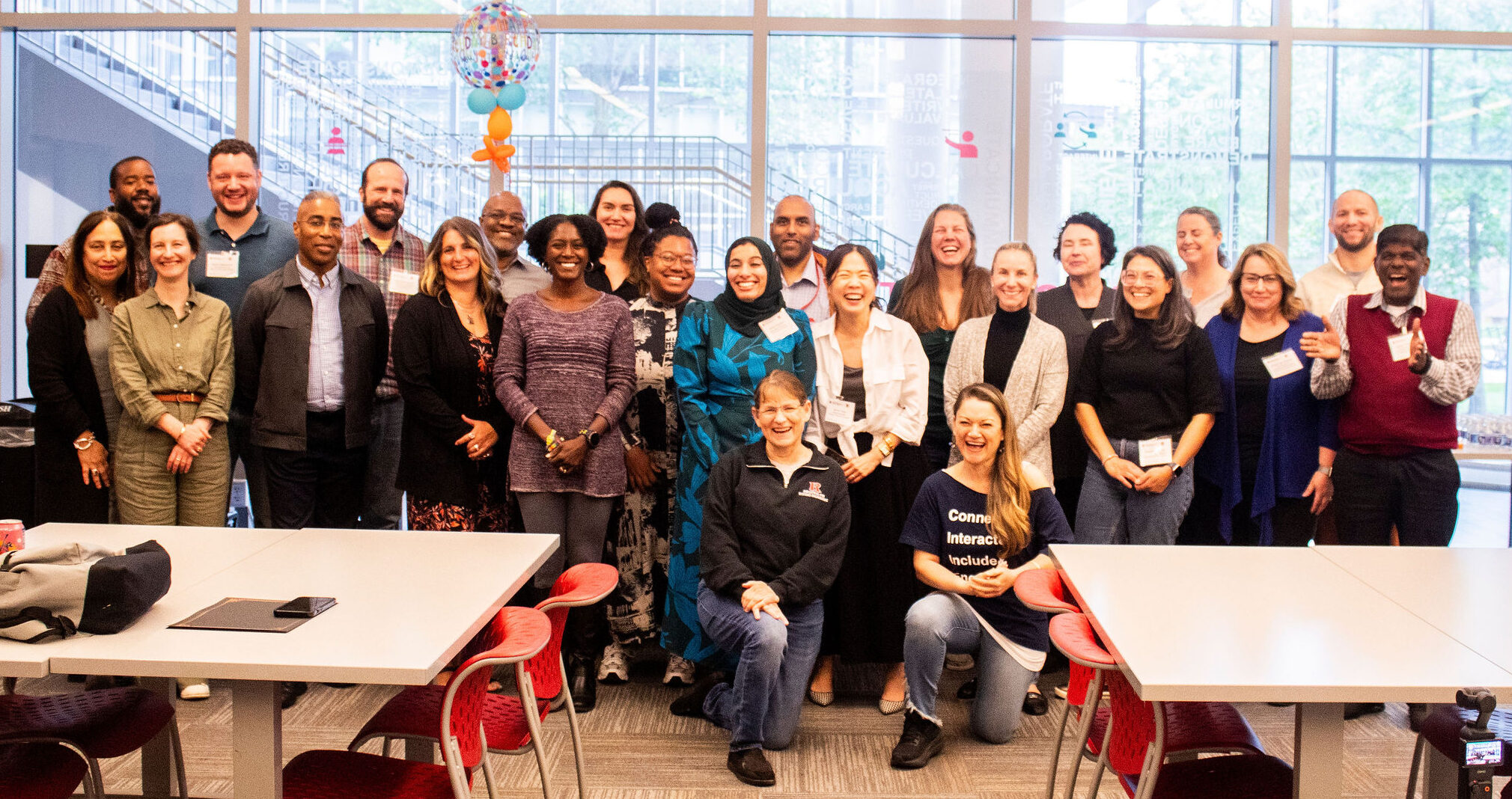 Group photo of approximately twenty participants gathered in a modern classroom or workshop space with large windows, red chairs, and a celebratory balloon visible above the group.