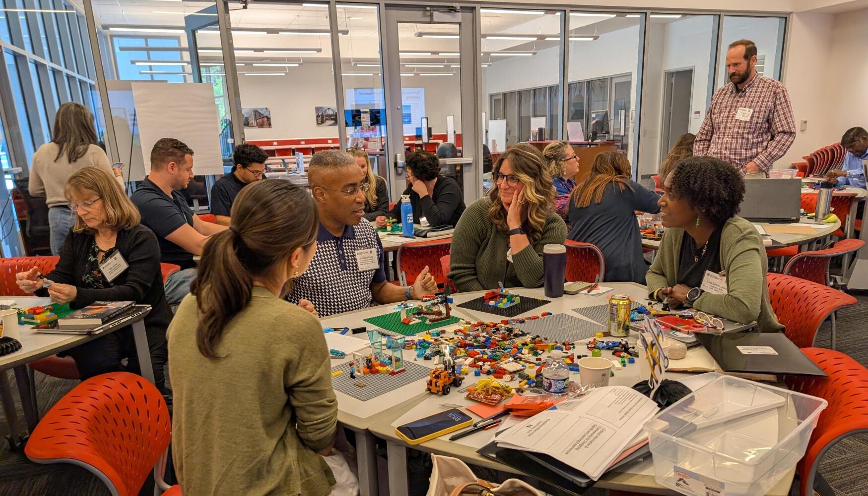 Workshop participants collaborate in small groups around tables, using colorful LEGO pieces and discussion materials in a bright, modern classroom.
