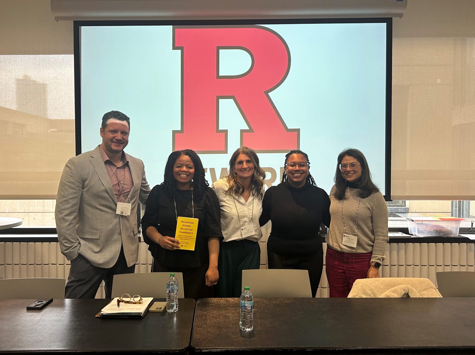Five participants pose for a photo in front of a projection screen displaying the Rutgers logo, behind conference tables set up for a workshop or panel discussion.