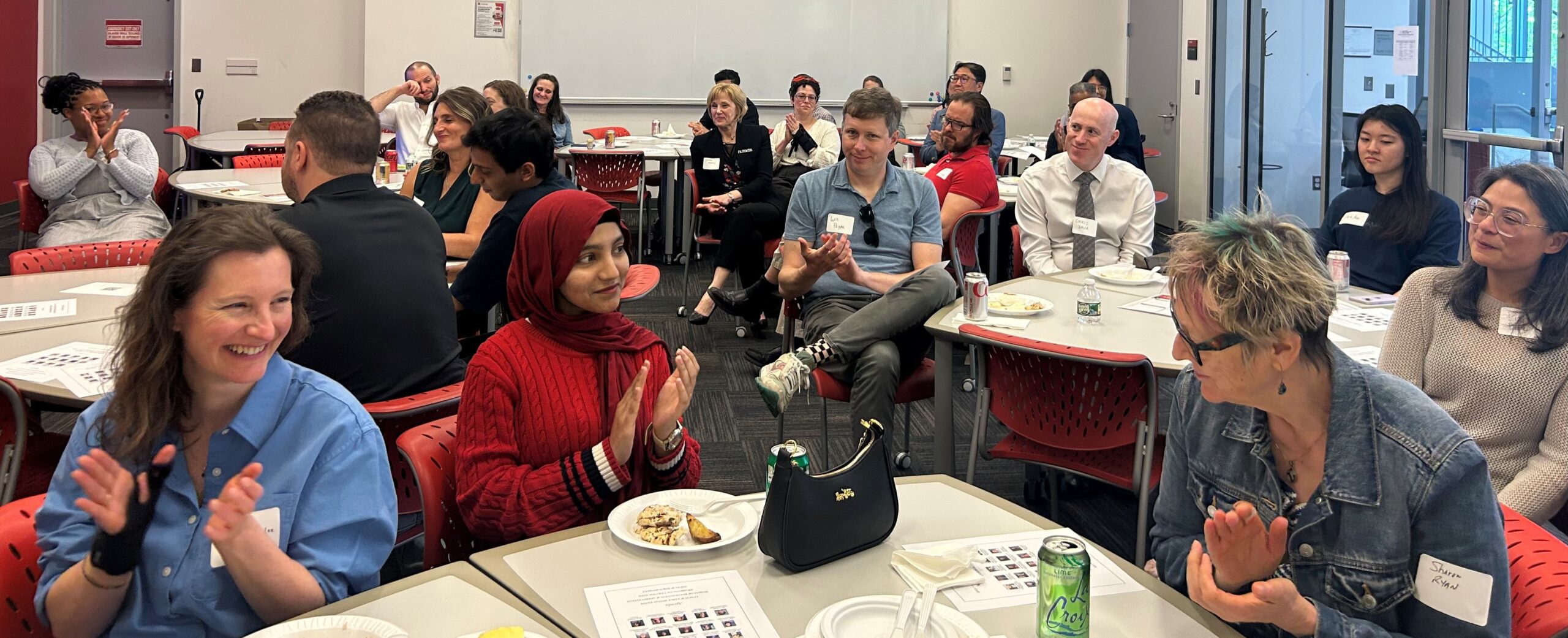 Workshop participants seated at round tables applaud during a group discussion or program session in a modern classroom.