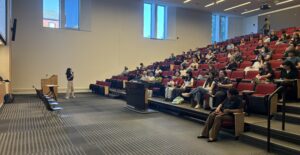 Lecture hall with tiered red seats filled with people listening to a speaker standing at the front near a podium and holding a microphone. Large windows provide natural light, and a ceiling‑mounted projector is visible above the audience.